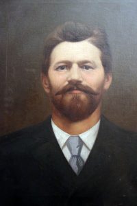 Porträt eines Mannes im dunklen Anzug, weißem Hemd und silbergrauer Krawatte. Er blickt ernst und hat braunes har und einen braunen Vollbart mit Schnurrbart.