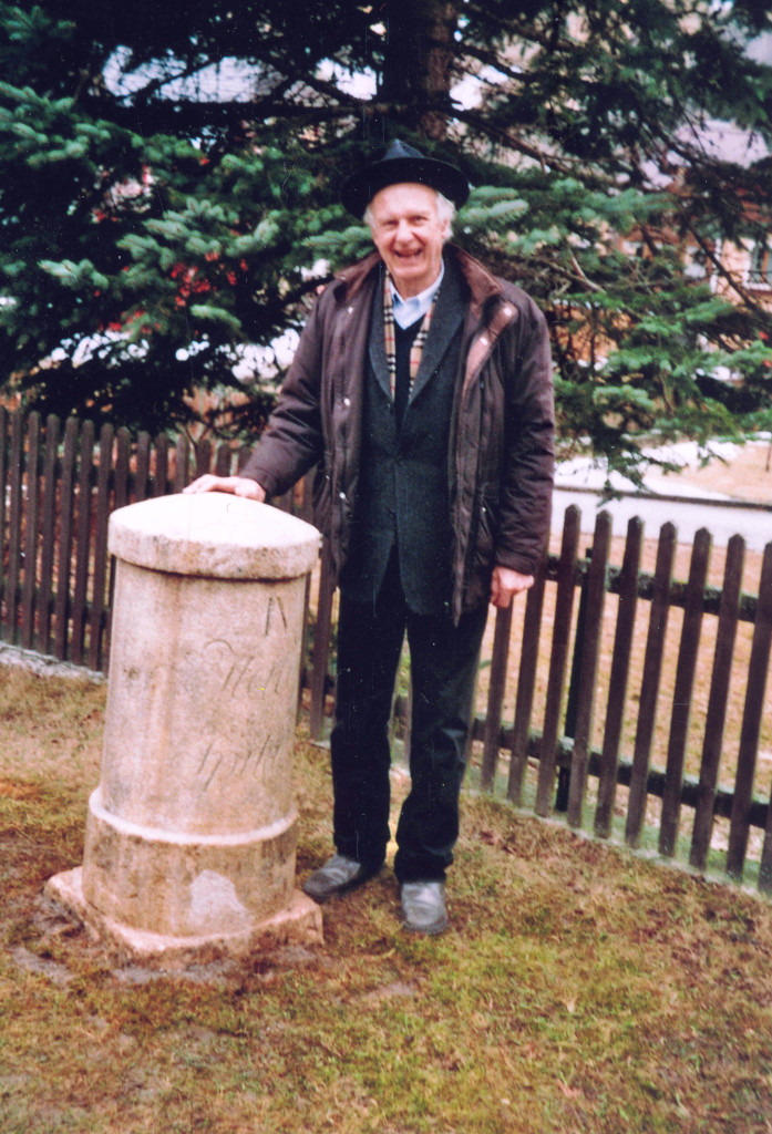 Ein älterer Herr mit schwarzem Hut, offener Winterjacke, schwarzem Sakko und schwarzer Hose steht rechts neben einem zylinderförmigen Stein, der ungefähr halb so groß ist wie er. Er legt seine linke Hand darauf. Im Hintergrund ein Holzlattenzaun und ein Nadelbaum.