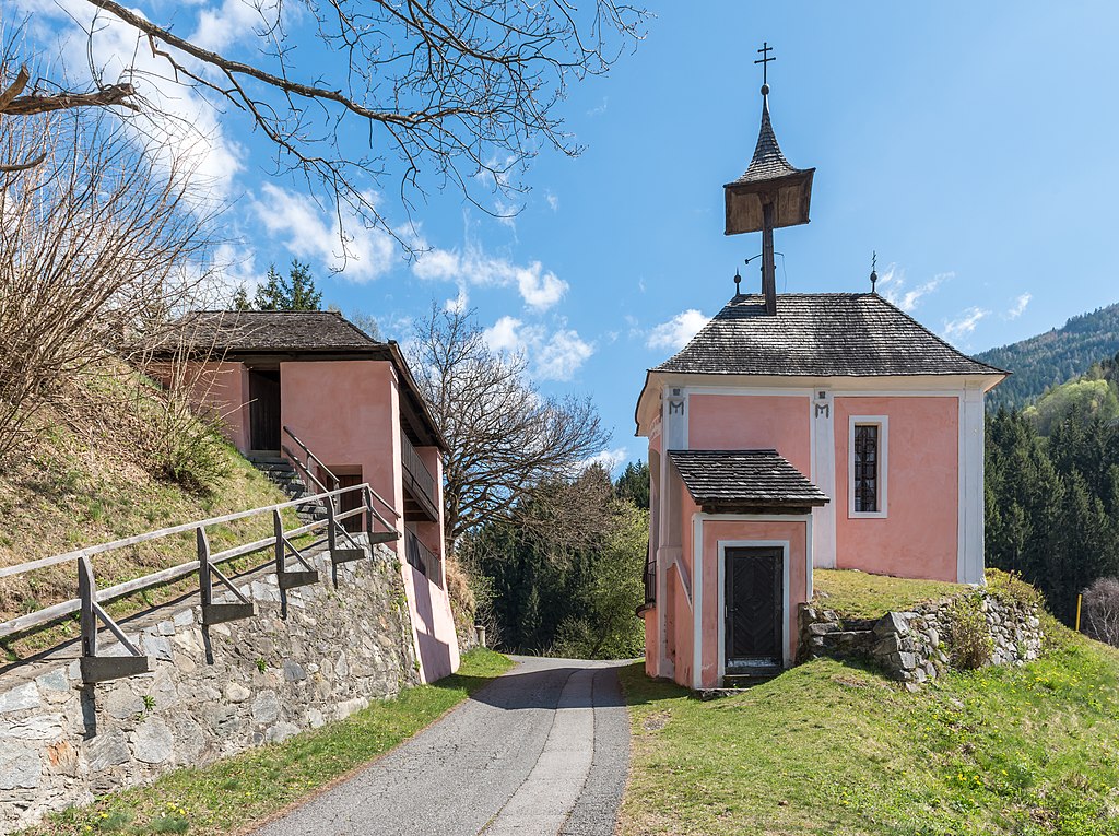 Auf einem Hügel eine darüberführende Straße und sowohl rechts als auch links davon ein rosa-farbenes Gebäude. Das Gebäude rechts davon ist als Kirchenbau zu erkennen.